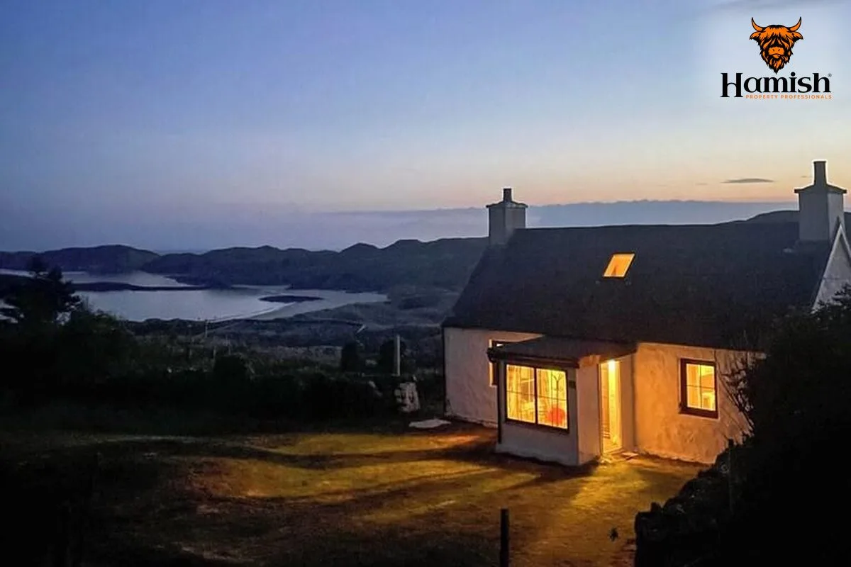 Stone Highland cottage with warm glowing windows overlooking a loch at sunset